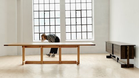Designer Stine Aas leaning over looking at the side of a Land Table in oak, with a Land Storage credenza against a wall in an open room.