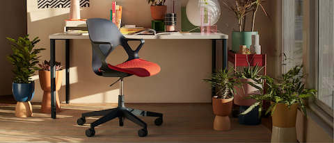 A dark Zeph Chair with a bright red 3D Knit seat pad sits in a sunny, plant-filled home office.