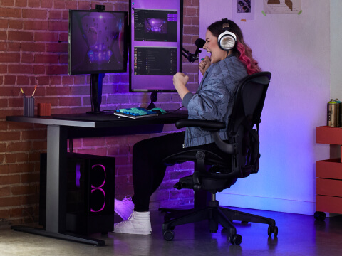A pink-haired woman cheers while sitting at her gaming setup with large monitors, sit-to-stand desk, and the Aeron Gaming Chair. A pink-haired woman cheers while sitting at her gaming setup with large monitors, sit-to-stand desk, and the Aeron Gaming Chair.